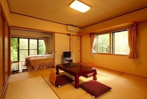 Ein traditioneller japanischer Raum mit Tatami-Bodenbelag, einem Bett, einem Fernseher und einem Tisch mit Kissen.