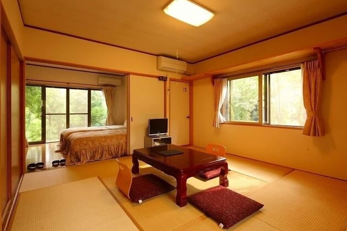 Ein traditioneller japanischer Raum mit Tatami-Bodenbelag, einem Bett, einem Fernseher und einem Tisch mit Kissen.