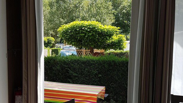 A room with a table and chairs, a view of a green landscape with trees and a hill, and a door with curtains.