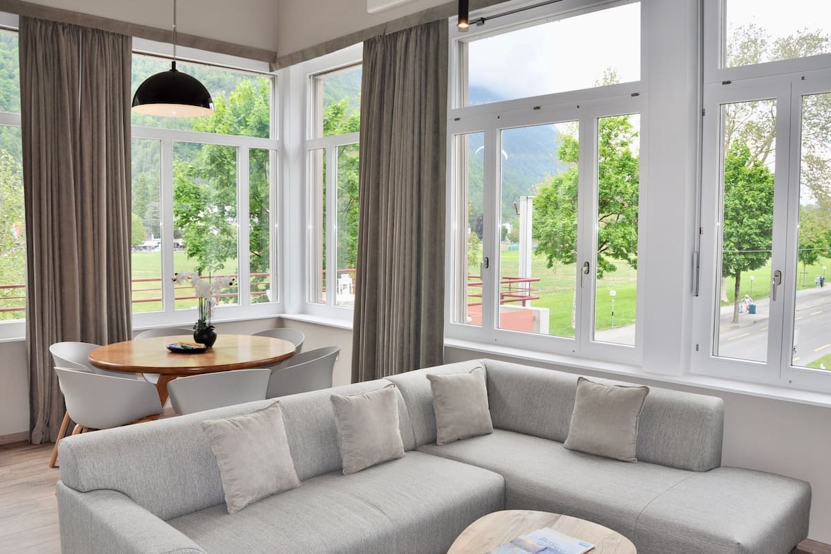 A modern living room with a grey sectional sofa, a round wooden coffee table, and a dining area with a white table and chairs. Large windows offer a view of a green park and mountains.