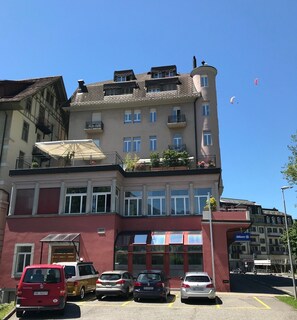 Hotel com vários andares, fachada em vermelho e bege, múltiplas varandas e área de estacionamento com vários carros.