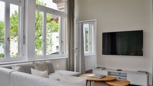 A modern living room with a large window, a flat-screen TV, a sofa, a coffee table, and a TV stand. - The Loft Interlaken (Interlaken)