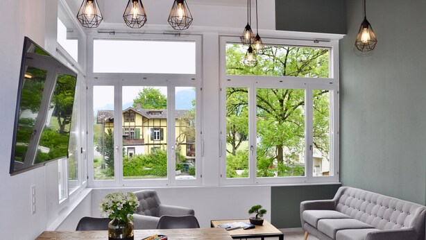 A modern living room with a dining area, large windows, and a grey sofa. - The Loft Interlaken (Interlaken)
