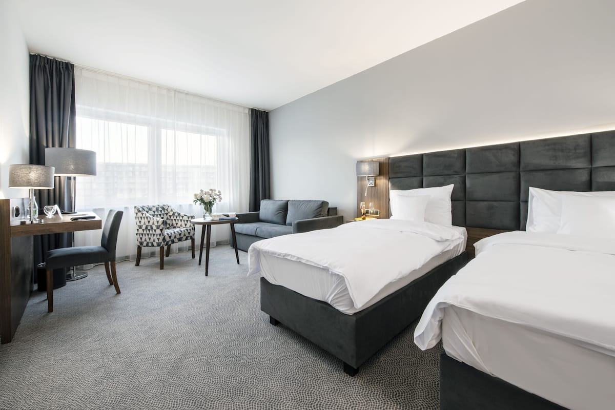 A hotel room with a large bed, a desk, a chair, and a small table with a lamp.