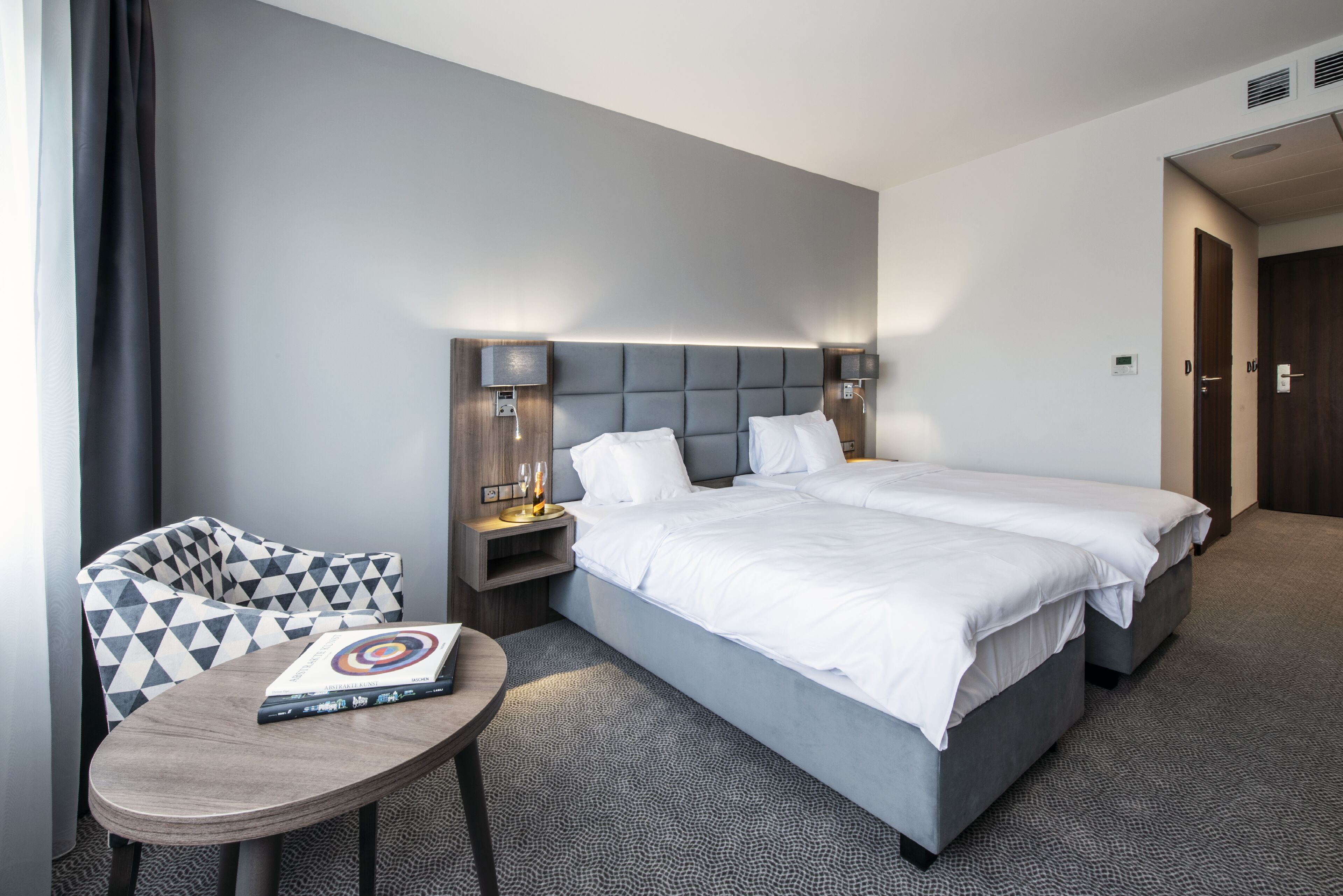 A modern hotel room with a large bed, a small table with a book, and a chair with a geometric pattern.