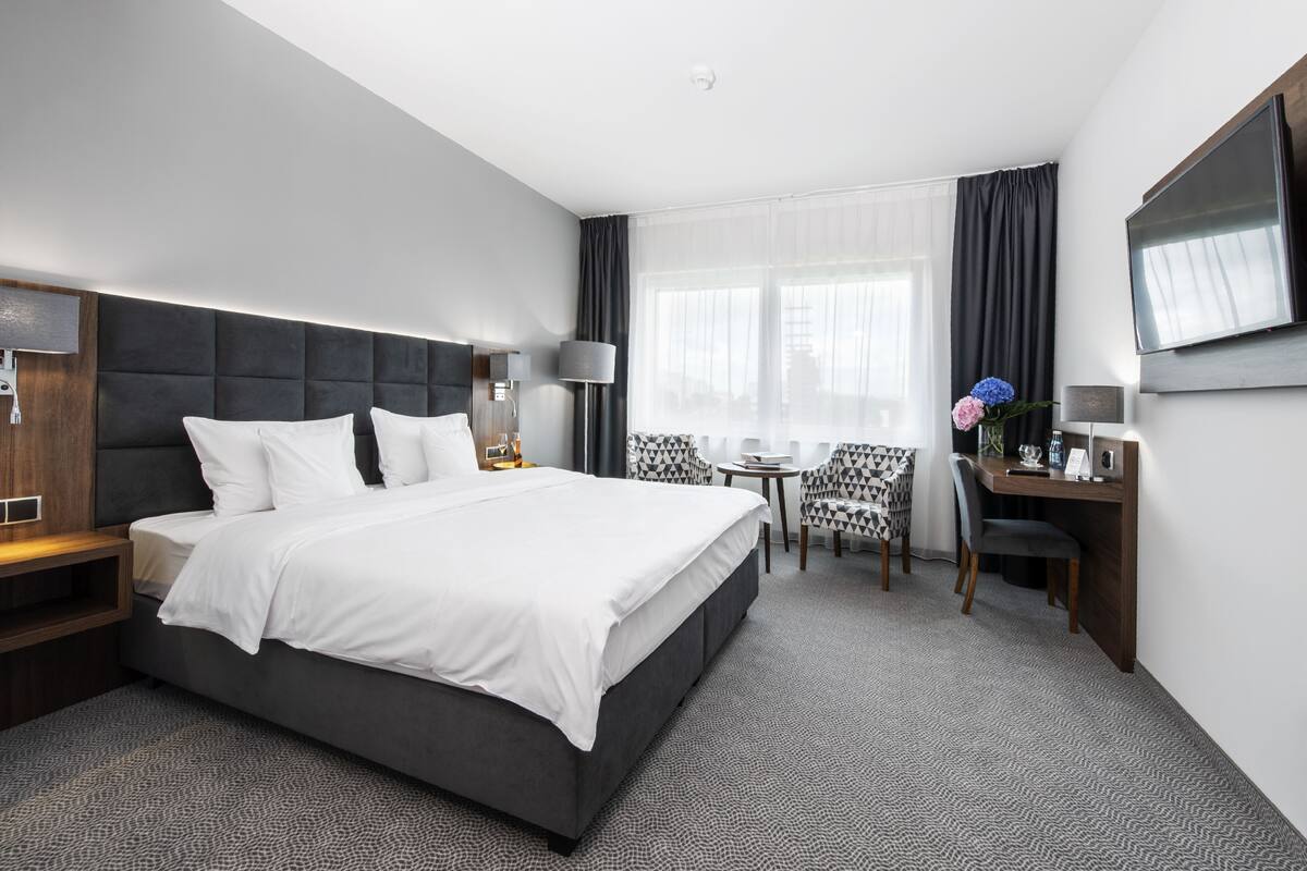 A modern hotel room with a large bed, a desk, a chair, a TV, and a window with curtains.