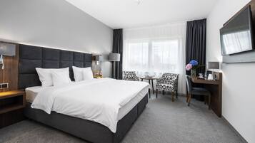 A modern hotel room with a large bed, a desk, a chair, a TV, and a window with curtains.