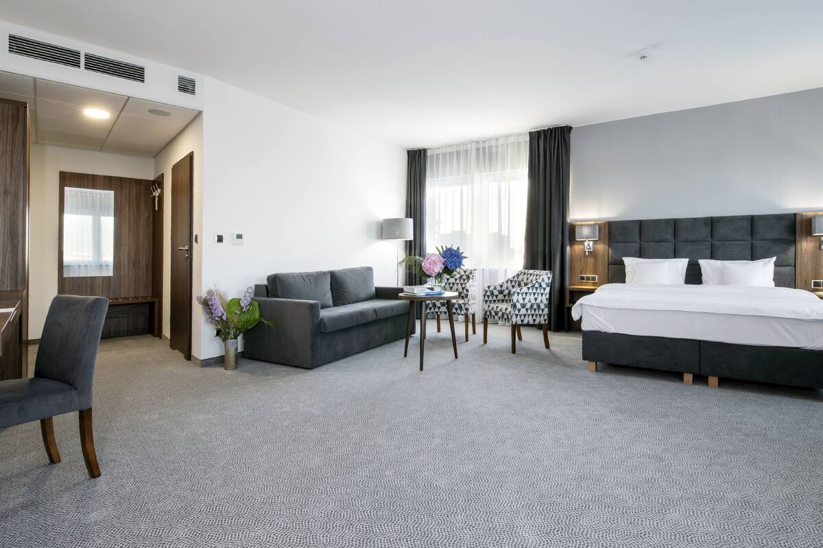 A hotel room with a bed, a sofa, a chair, a small table, and a lamp.
