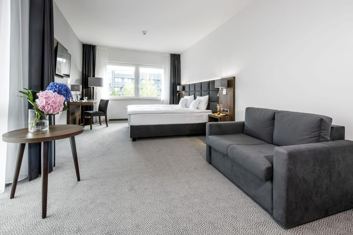 A modern hotel room with a bed, a gray sofa, a small table with a vase of flowers, a desk with a chair, and a TV mounted on the wall.