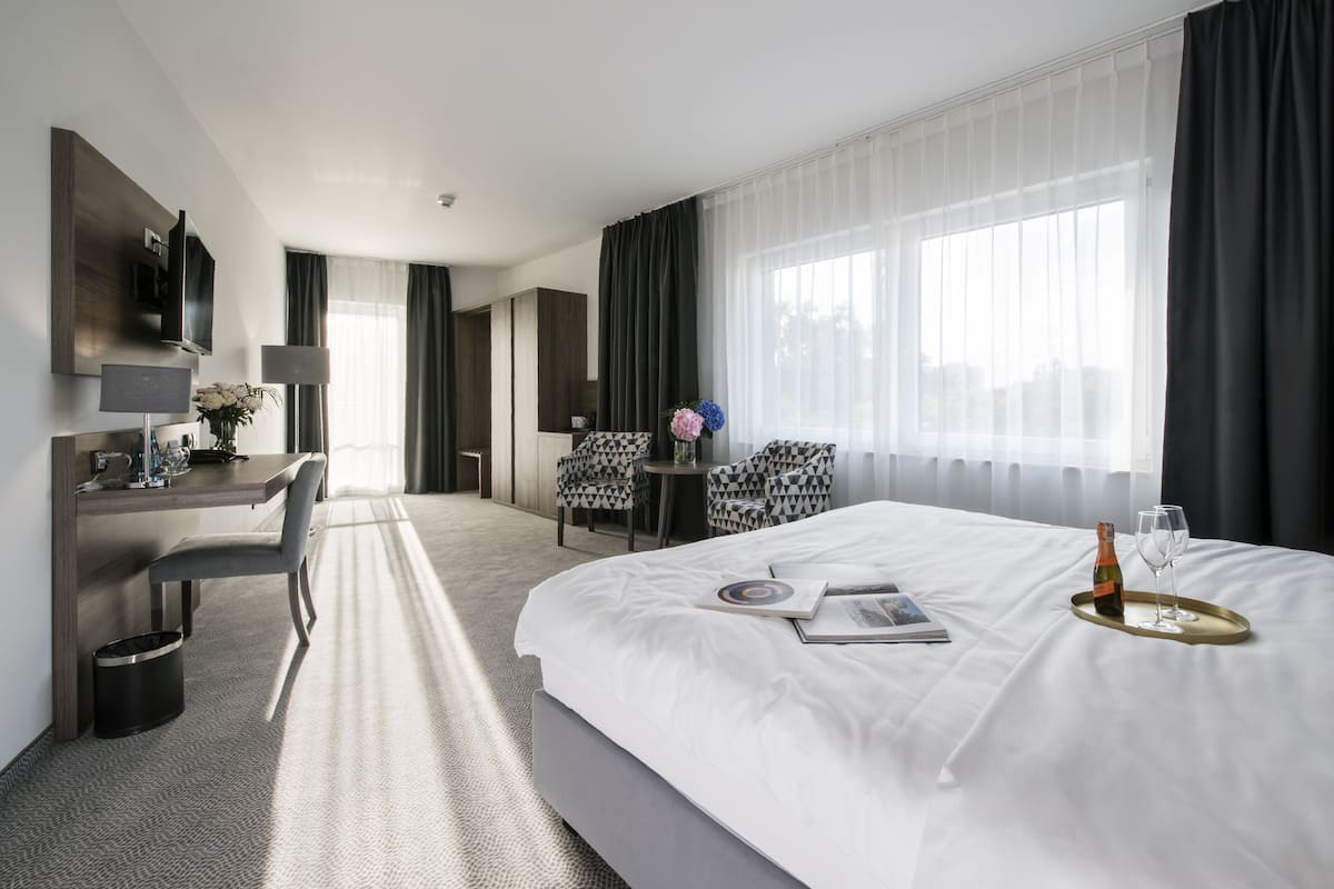 A modern hotel room with a large bed, a desk with a chair, a TV, and a view of the city through the window.