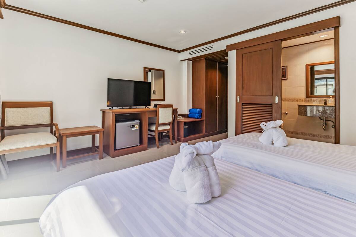 A hotel room with two beds, a desk with a television, a small refrigerator, and a wardrobe.