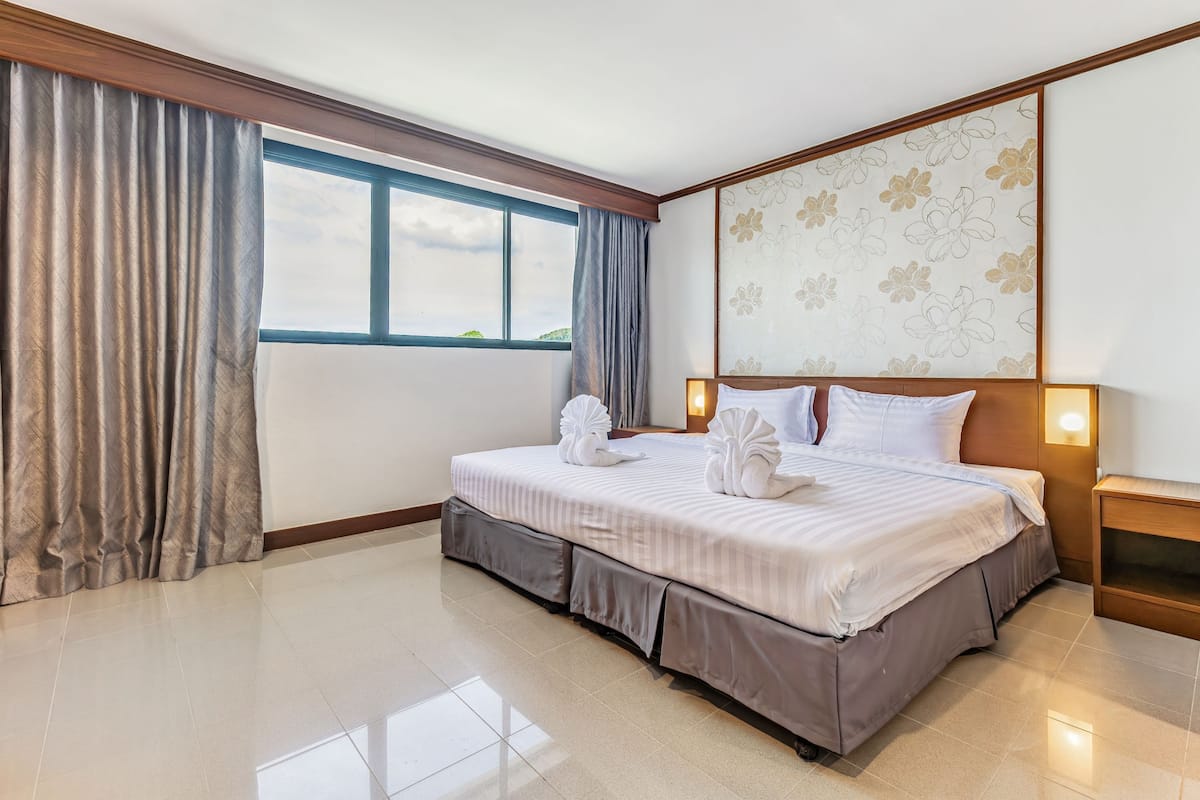 A hotel room with a large bed, a window with curtains, a floral wall decoration, and a nightstand with a lamp.