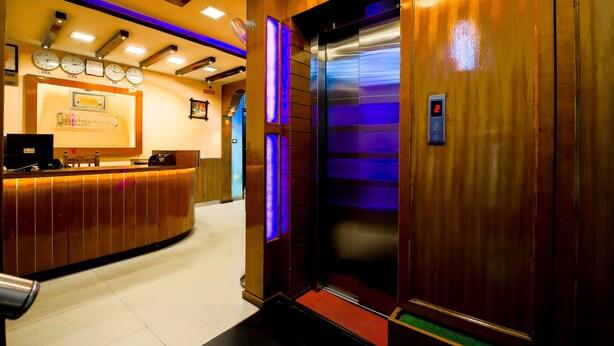 A hotel reception area with a wooden reception desk, a digital clock, and an elevator with a purple light. - Lusia Inn (Bengaluru)