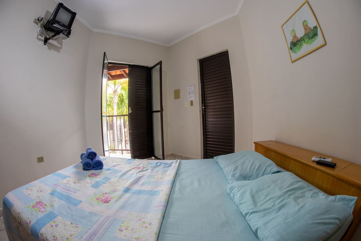 A bedroom with a bed, a wooden nightstand, a TV mounted on the wall, and a door leading to an outdoor area.