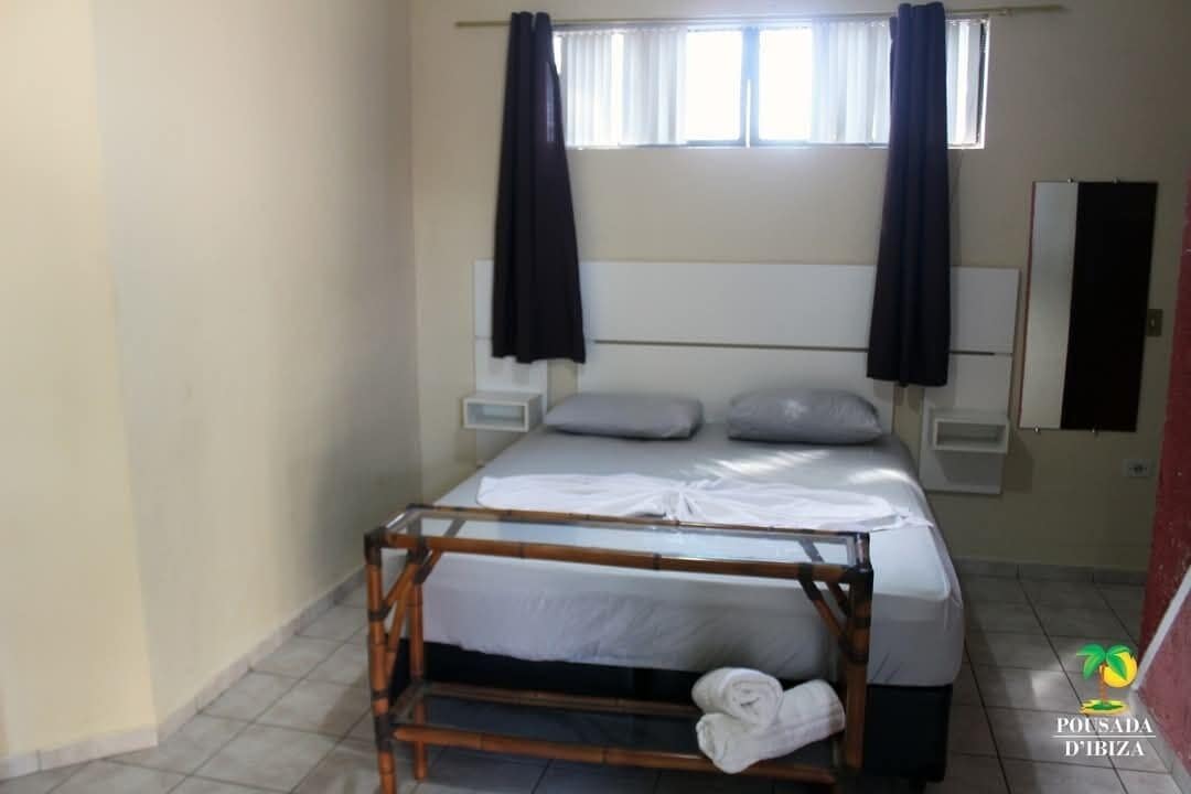A single-bed room with a wooden bunk bed, white bedding, a small bedside table, and a window with curtains.