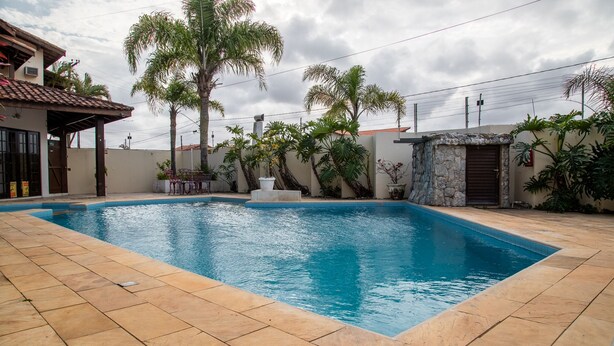 A rectangular swimming pool with a tiled deck, surrounded by palm trees and a stone wall. - Pousada D'Ibiza (Itanhaém)
