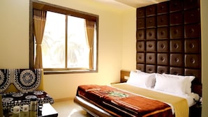 A hotel room with a bed, a ceiling fan, a window with curtains, a bedside table with a coffee maker, and a decorative headboard. - Hotel Shahi Darbar (Navsari)