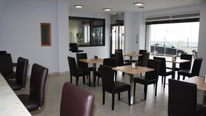 Een restaurant met meerdere tafels en stoelen, een raam met uitzicht op een auto buiten, en een deur met een nooduitgangsbord.