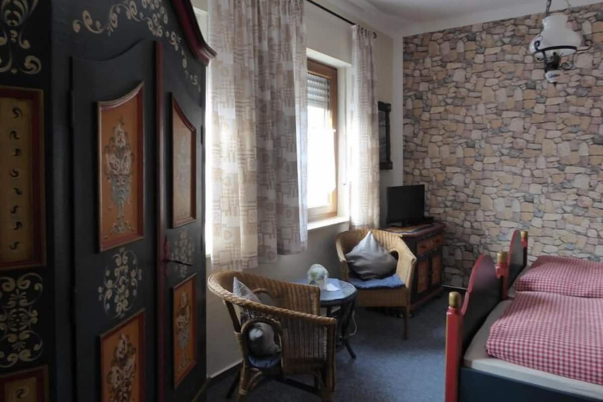 Una habitación acogedora con pared de piedra, una cama con cubrecama a cuadros, una silla de mimbre, una mesita con un florero y un televisor sobre un mueble de madera.