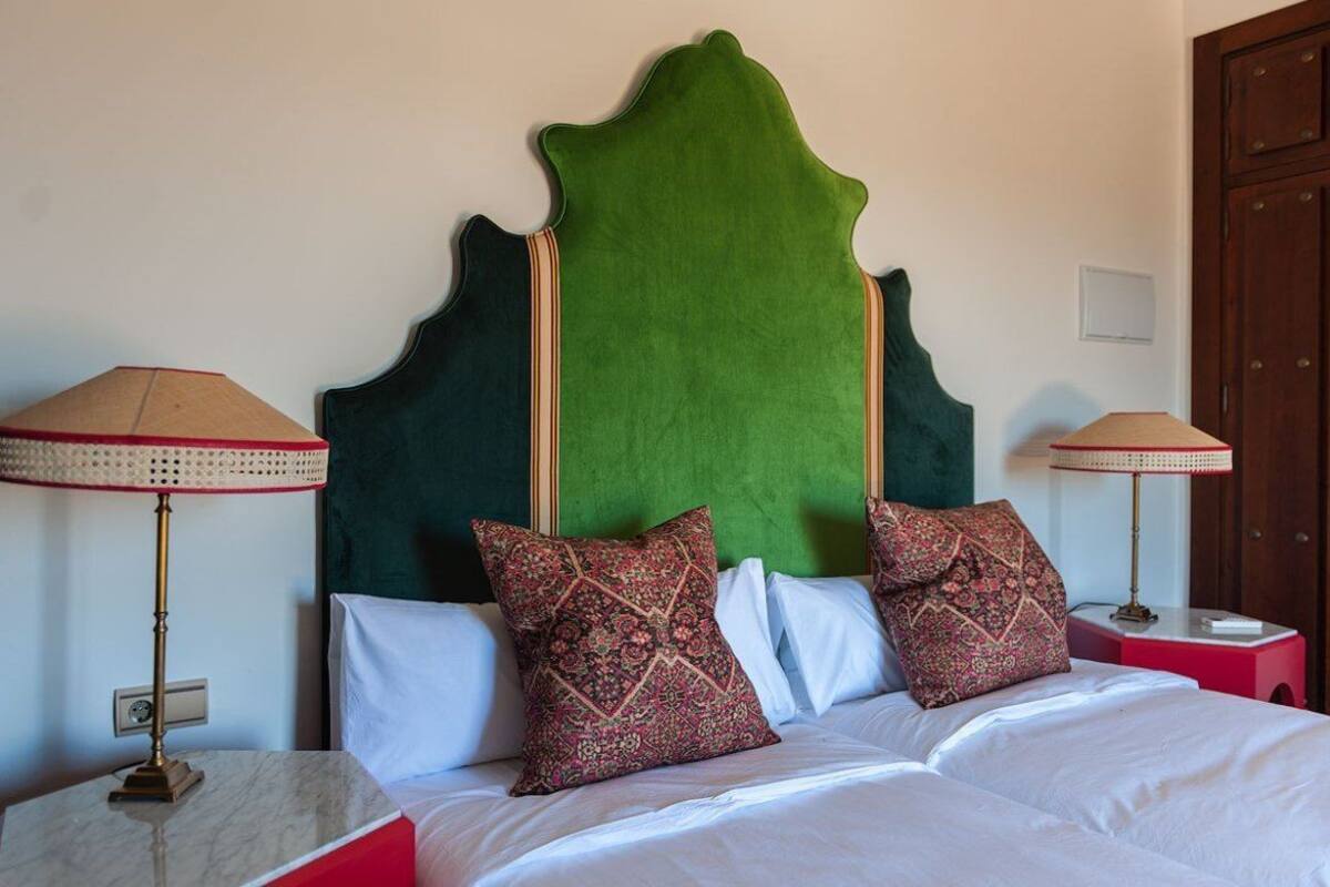 Ein Bett mit weißen Leinentüchern, zwei gemusterten Kissen, einem grünen gepolsterten Kopfteil und zwei Nachttischlampen mit beige farbenen Lampenschirmen.