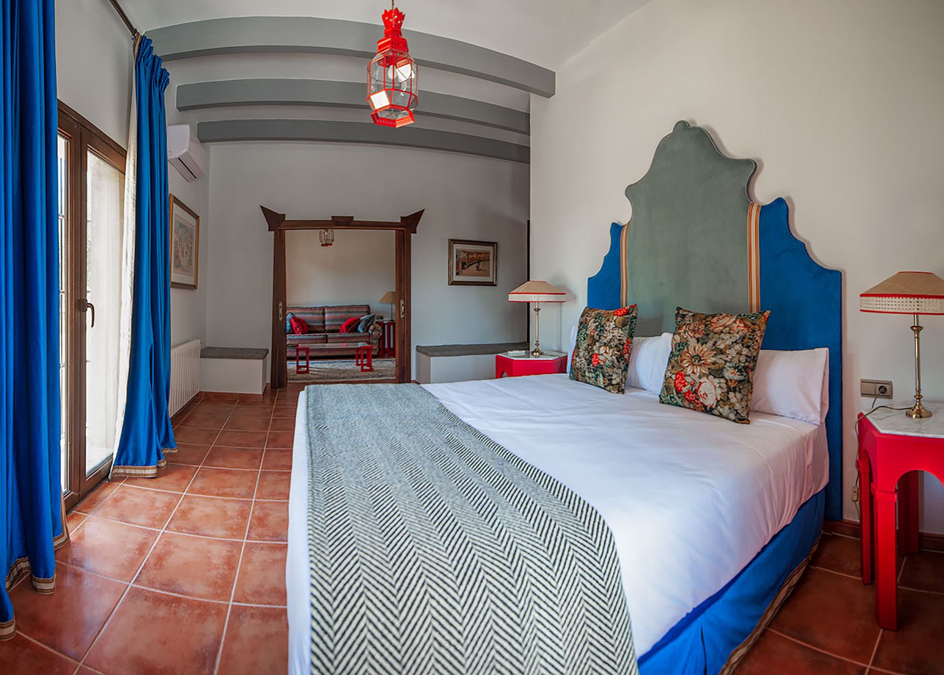 Une chambre d’hôtel avec un grand lit, une table de chevet rouge, une tête de lit bleue et une vue sur l’espace extérieur.