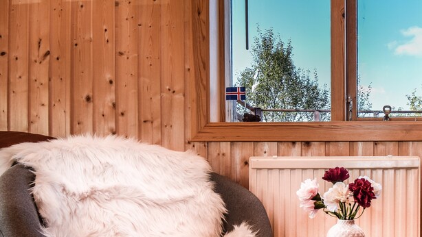 Een gezellige kamer met een houten wand, een raam met uitzicht, een zacht wit kussen en een vaas met bloemen.