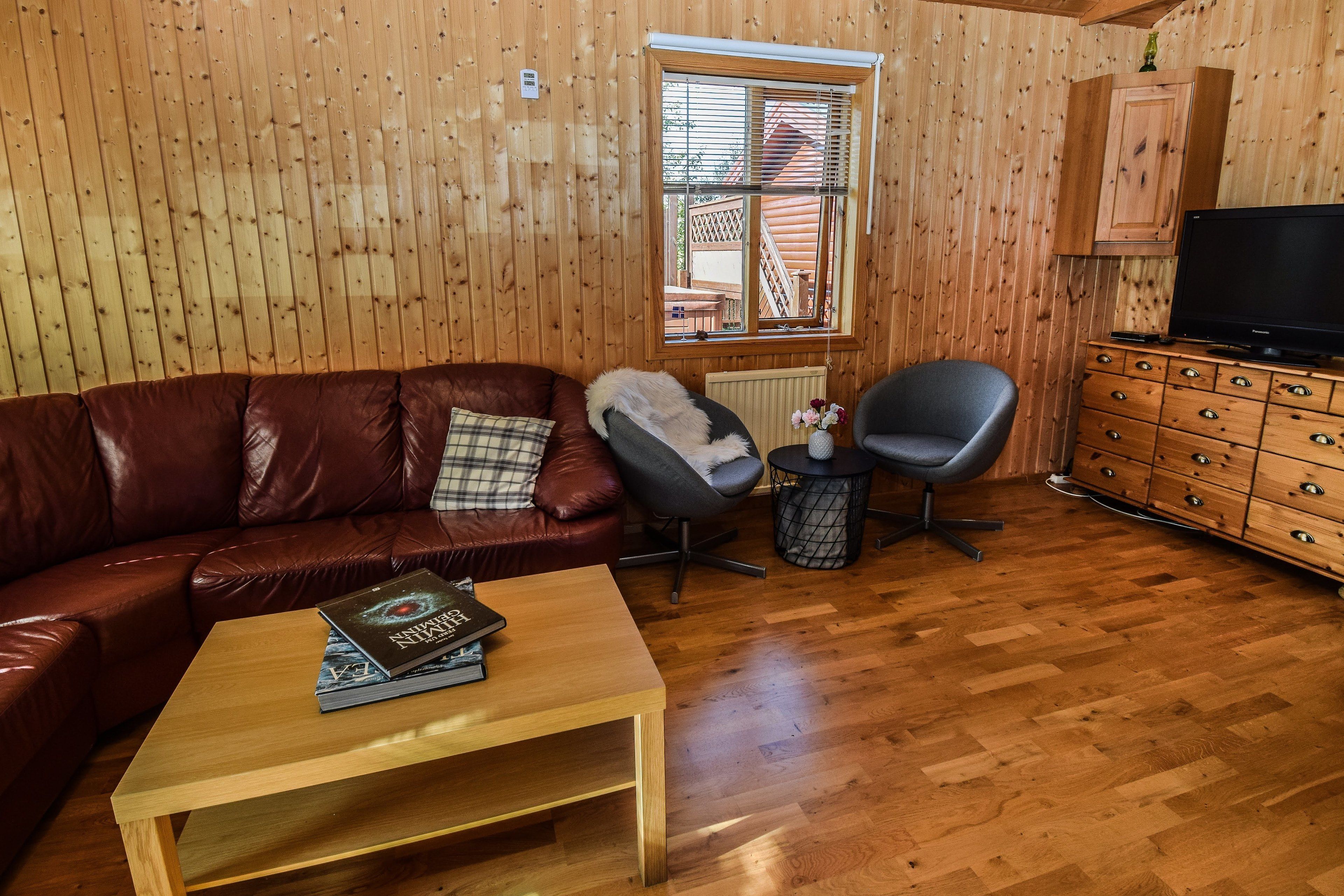 Una habitación con paneles de madera, un sofá de cuero, una pequeña mesa con libros, un sillón gris, un aparador de madera y una televisión.