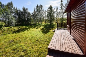 A wooden deck with a railing, surrounded by a grassy area and trees. - Cottage O12 (Selfoss)