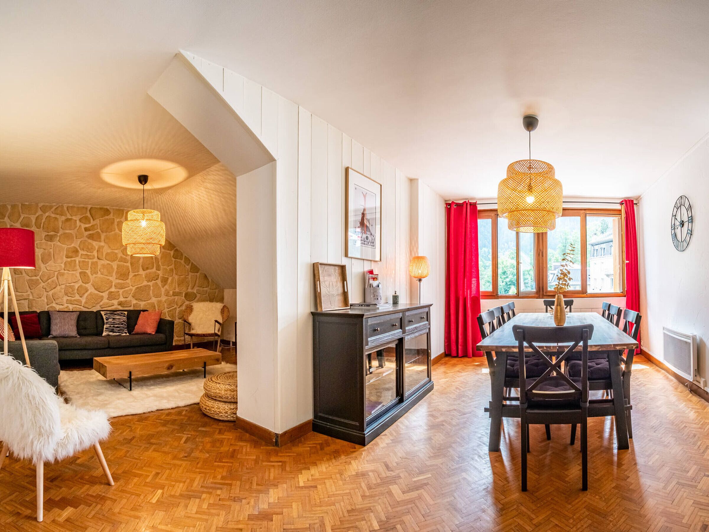 Ein gemütliches Wohnzimmer mit Steinwand, Kamin, Essbereich mit Holztisch und Stühlen sowie einem großen Fenster mit roten Vorhängen.