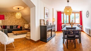 A cozy living room with a stone wall, a fireplace, a dining area with a wooden table and chairs, and a large window with red curtains. - Stunning downtown apartment - Pets allowed, WiFi (Chamonix-Mont-Blanc)