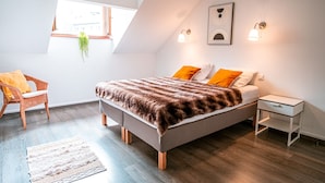 A modern bedroom with a loft-style ceiling, a bed with a fur throw, two orange pillows, a white nightstand, and a wicker chair. - Stunning downtown apartment - Pets allowed, WiFi (Chamonix-Mont-Blanc)