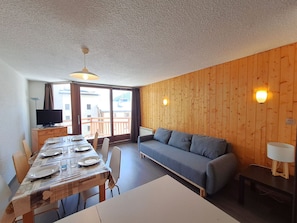 Une pièce avec un mur en bois, un canapé, une table à manger avec des chaises, un téléviseur et un balcon.