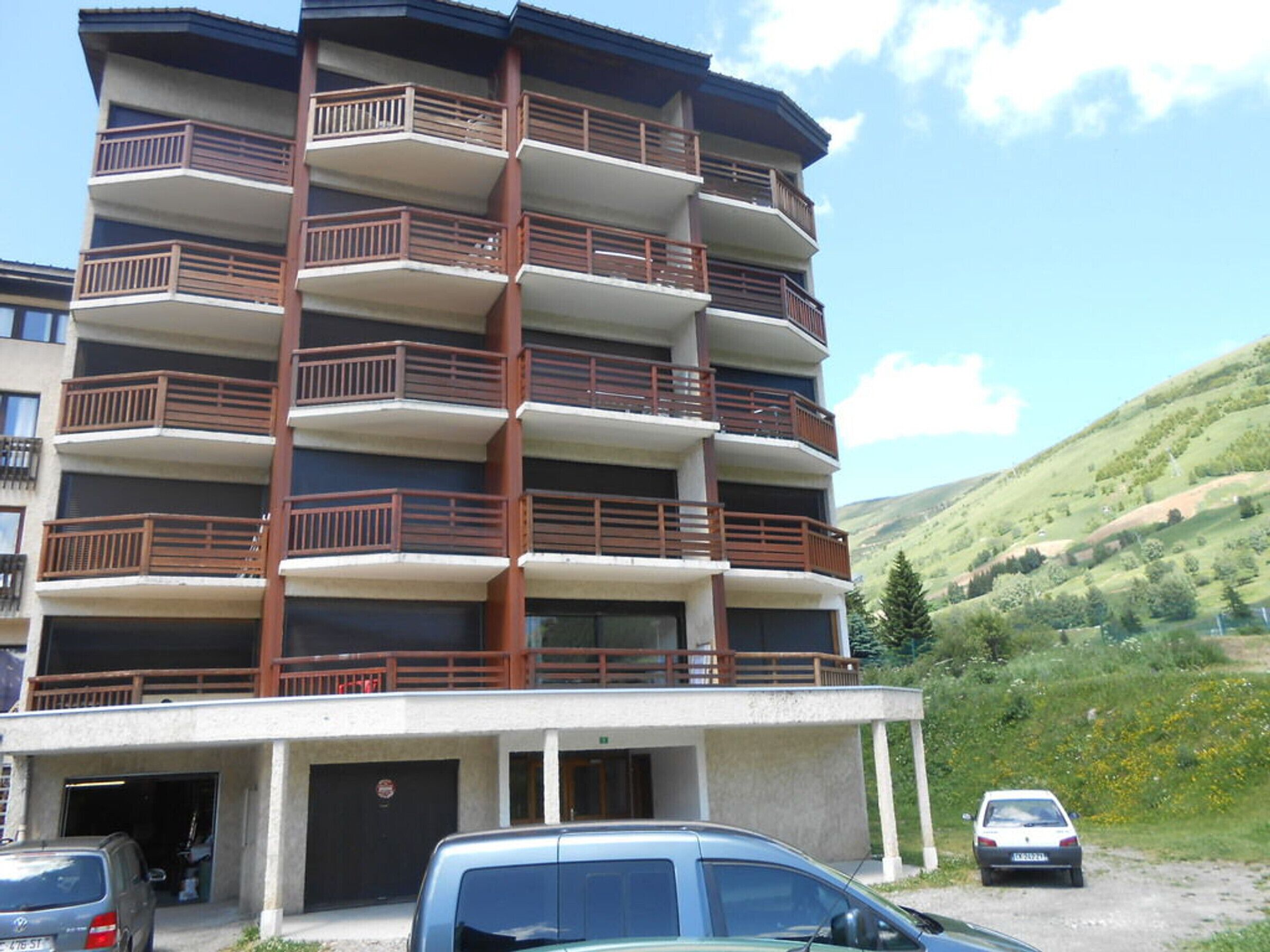 A multi-story building with balconies, a parking area, and a hilly landscape in the background.