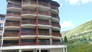 A multi-story building with balconies, a parking area, and a hilly landscape in the background.