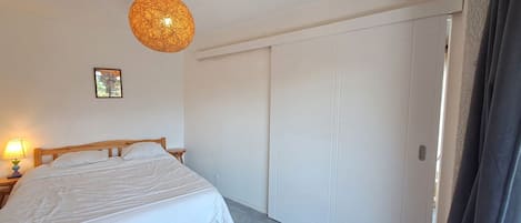 A bedroom with a bed, a hanging light fixture, a door, and a small table with a lamp.