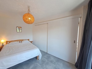 A bedroom with a bed, a hanging light fixture, a door, and a small table with a lamp.