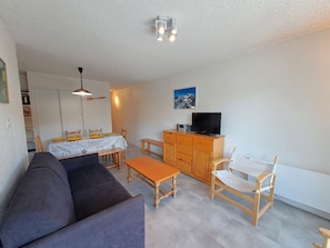 Ein Wohnzimmer mit einer Couch, einem Esstisch mit Stühlen, einer Holzkommode mit Fernseher und einer Deckenleuchte.