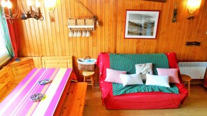 Interior acogedor de una cabaña con una pared de madera, un sofá rojo con cojines, una mesa a rayas y una lámpara colgante.
