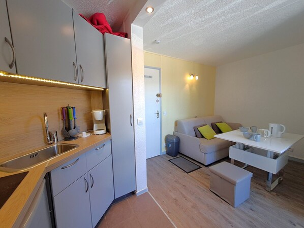 A compact kitchenette with a sink, a coffee maker, and a small countertop. There is a sofa and a table with cups and a kettle.