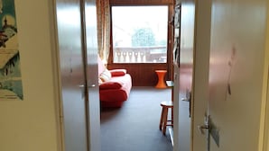 Un couloir étroit mène à une pièce où se trouvent un lit, une petite table et une fenêtre avec des rideaux.
