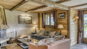 Una sala de estar acogedora con chimenea de piedra, vigas de madera, un conjunto de sofás y vistas al exterior a través de amplios ventanales.