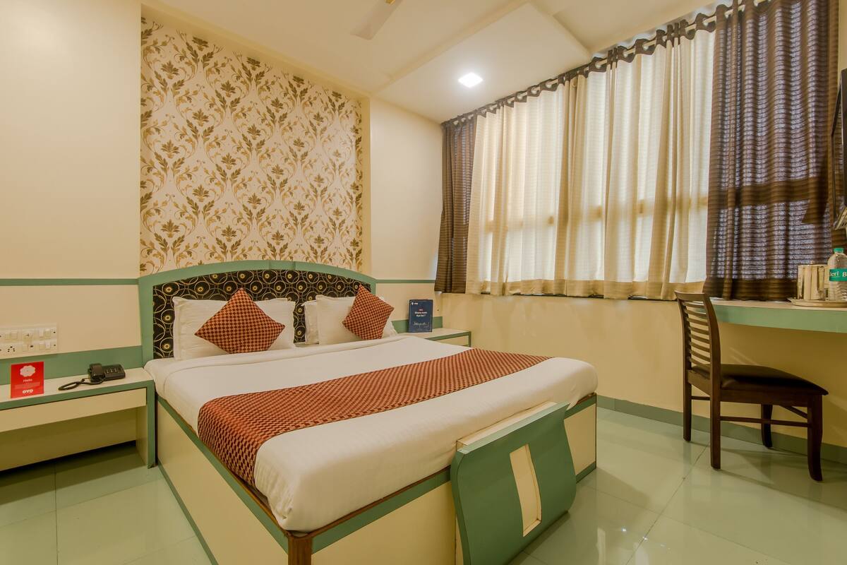 A hotel room with a bed, a desk, a chair, a nightstand, and a window with curtains.