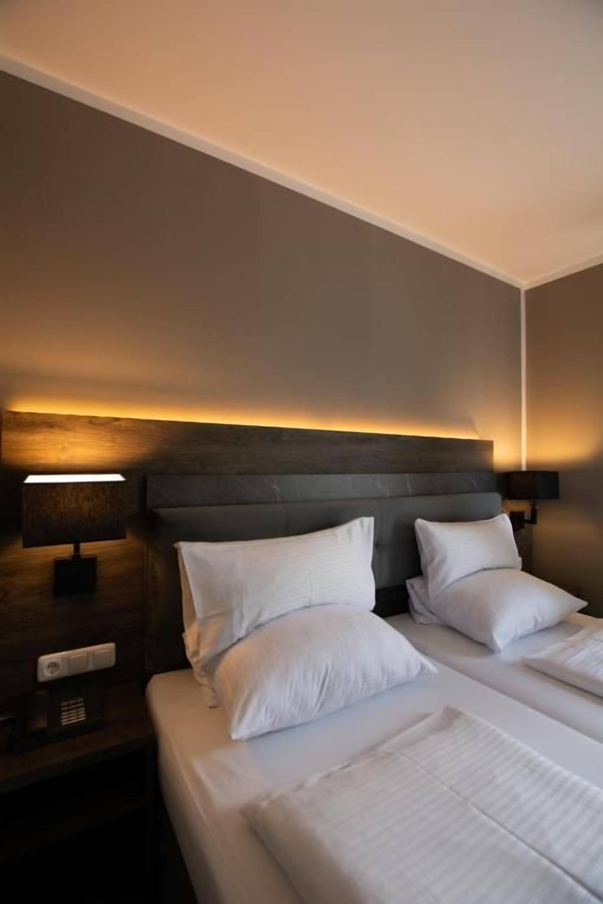 Habitación de hotel con cama, dos almohadas, mesita de noche y lámpara de pared.