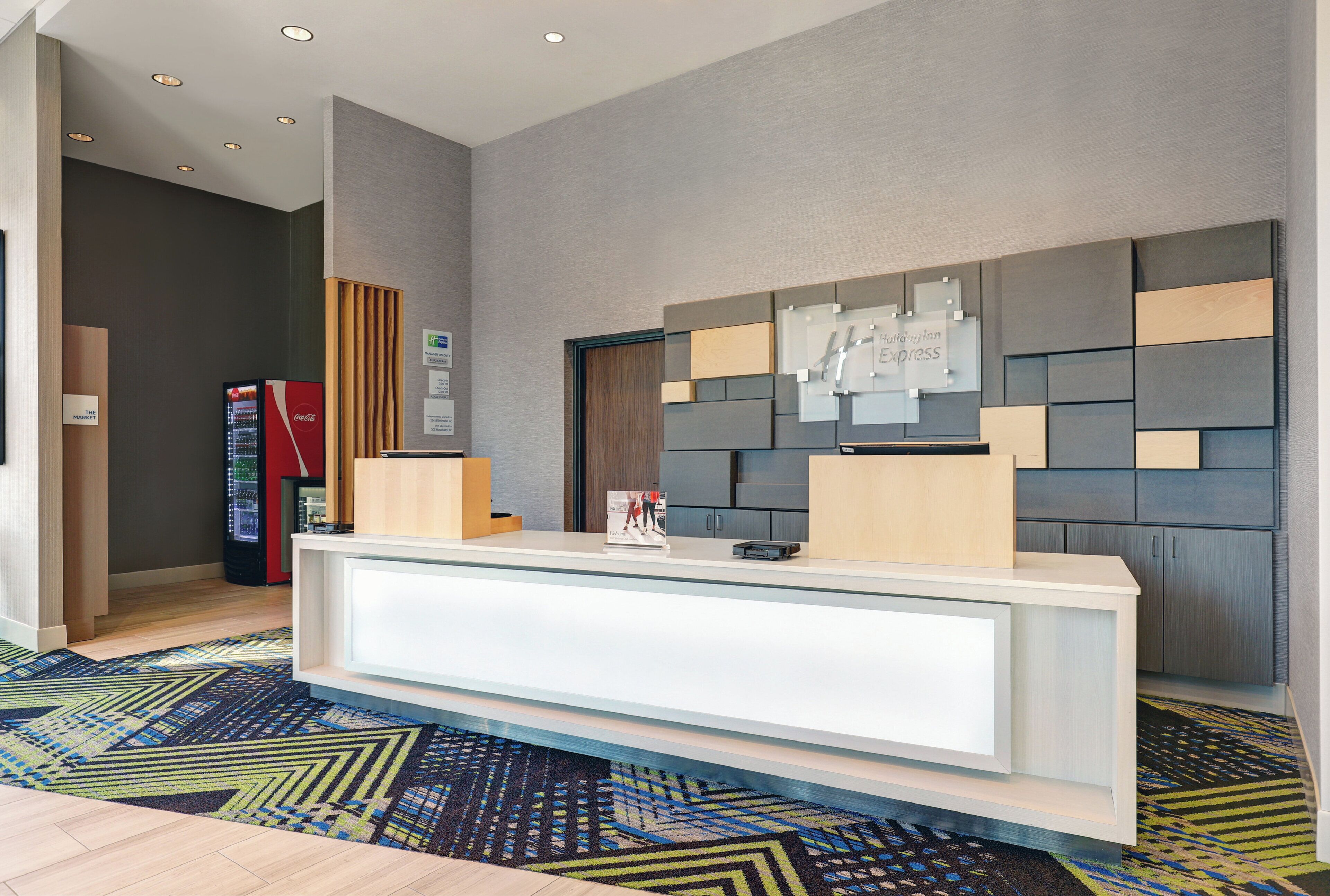A hotel reception area with a modern desk, a Coca-Cola refrigerator, and a patterned carpet.