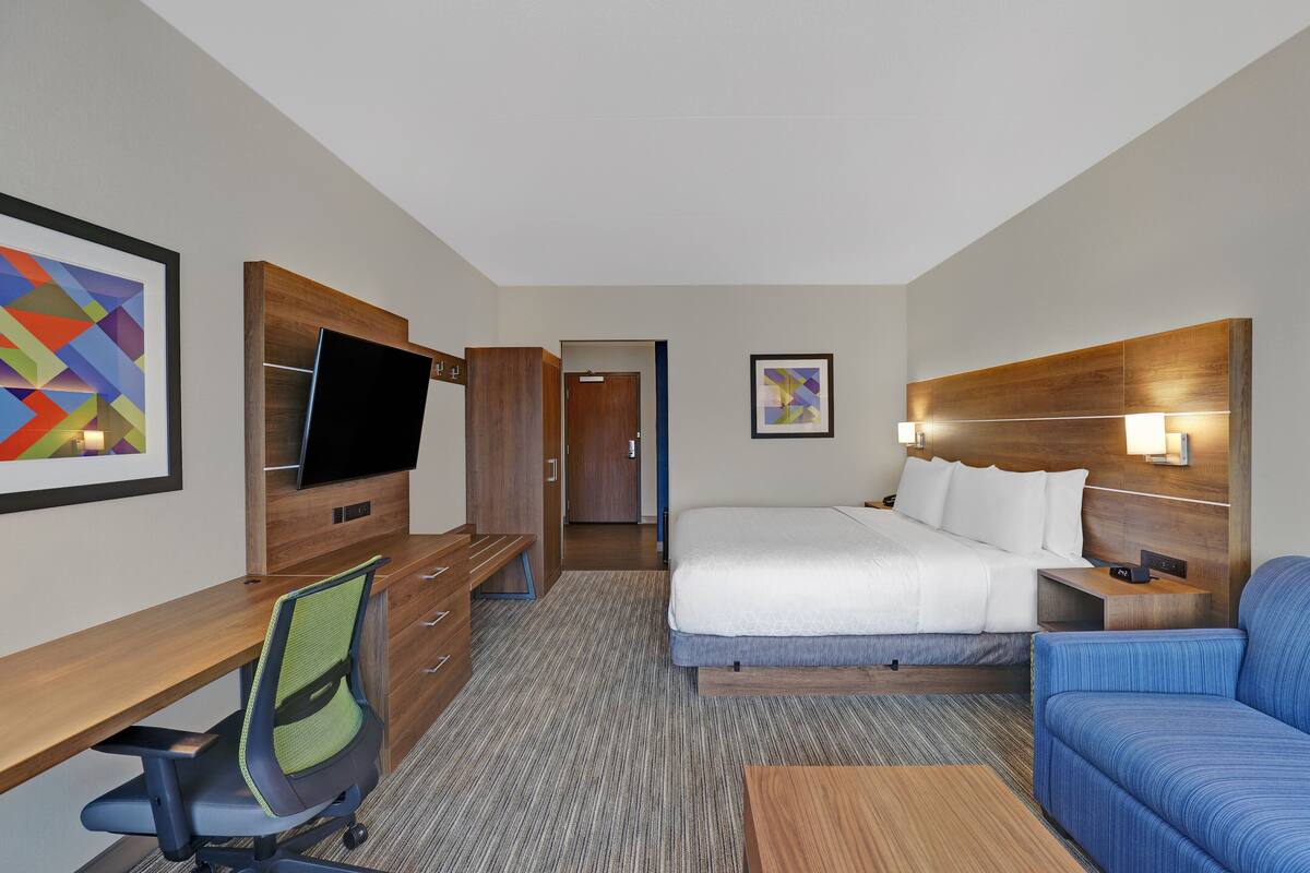 Una habitación de hotel con una cama, un escritorio con silla, un televisor, un sofá y obras de arte en las paredes.