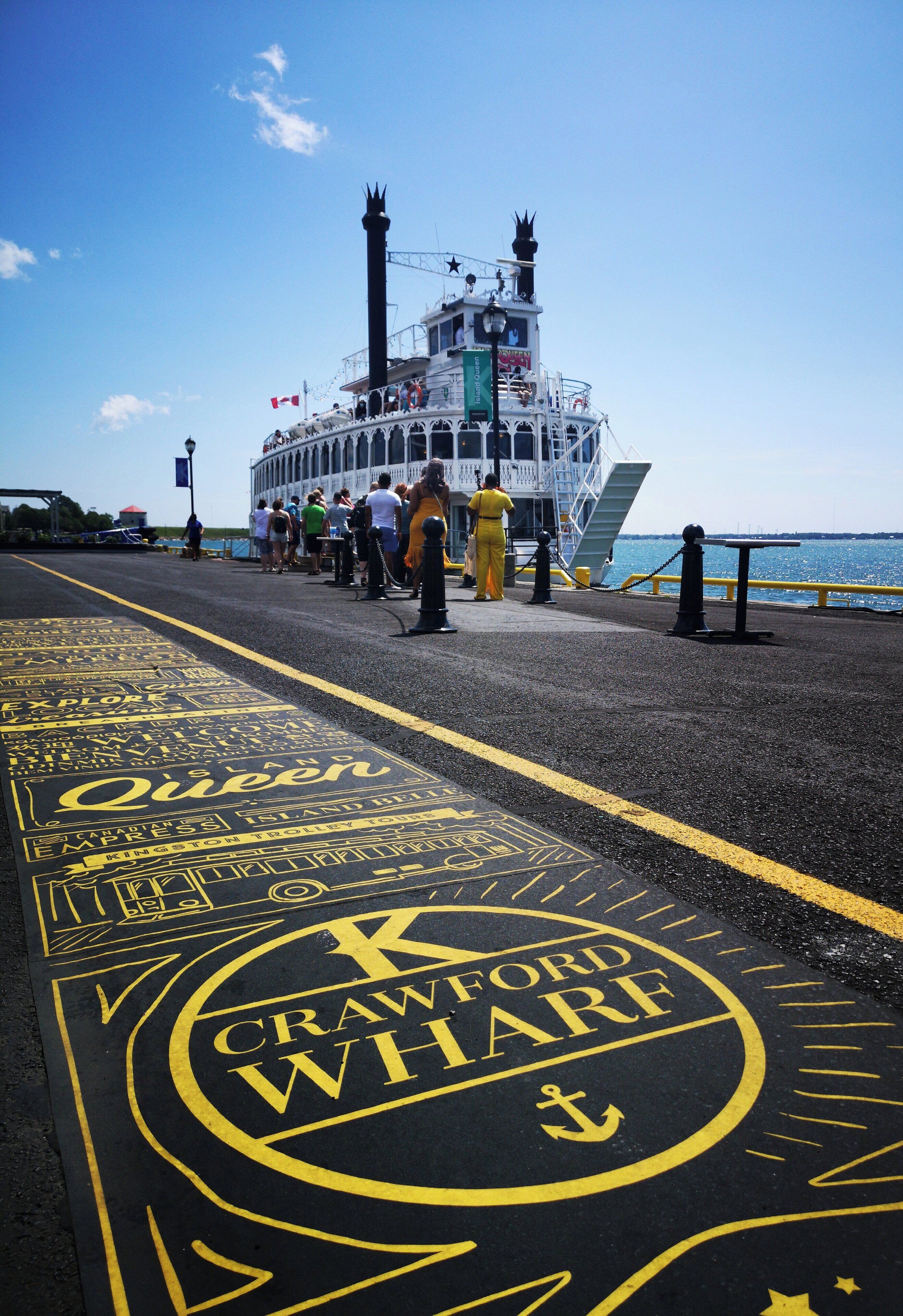 A ferry dock with a large ship in the background, a walkway with yellow markings, and people boarding the ferry.