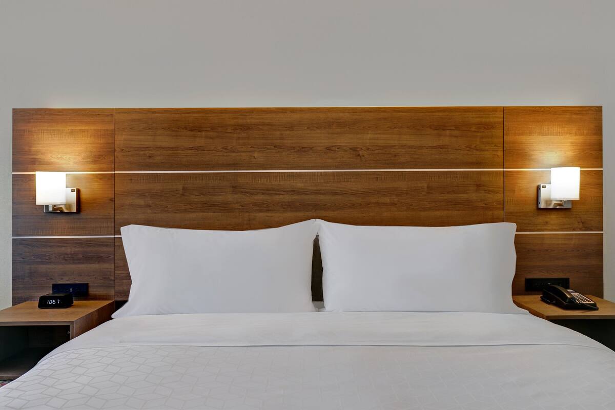 Habitación de hotel con cabecera de madera, dos lámparas de noche, cama con ropa blanca y mesita de noche con un teléfono y un reloj.