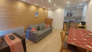 Ein gemütlicher Wohnbereich mit grauem Sofa, einer Holzwand, einem Esstisch mit rotem Tischläufer und einer Küche im Hintergrund.