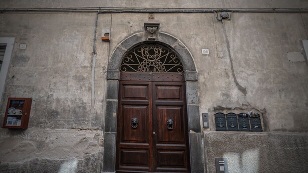 Una puerta doble grande de madera oscura con un arco de metal decorativo encima, ubicada en una pared de piedra desgastada con placas numeradas y un pequeño armario a la izquierda.