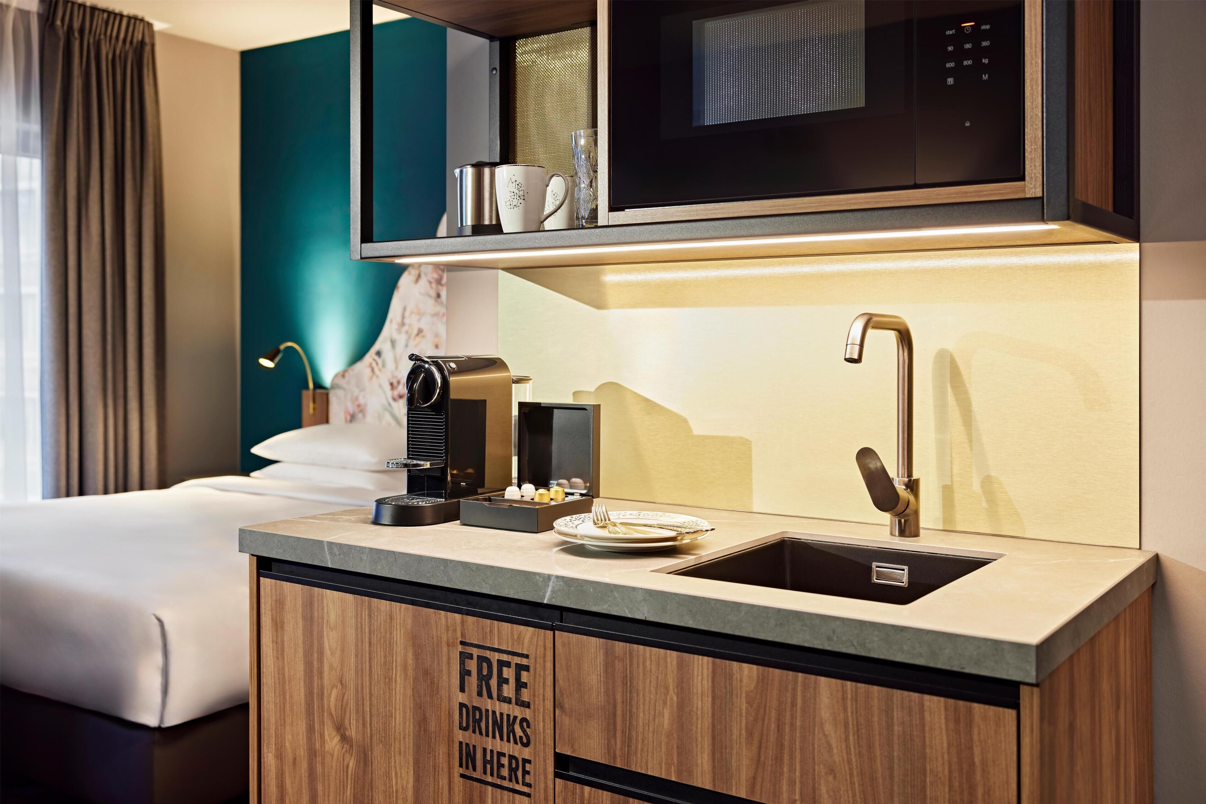 Habitación de hotel compacta con kitchenette, una cama, una cafetera y un cartel que indica bebidas gratuitas.